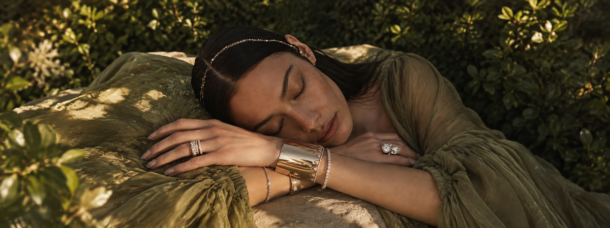 A woman lying down outdoors wearing gold jewelry, including a cuff bracelet, rings, and a headband, with green foliage in the background.